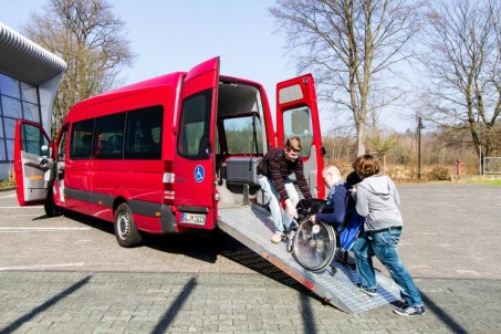 Das Foto zeigt einen roten Mercedes-Bus, in den Schüler über eine Rampe einsteigen.