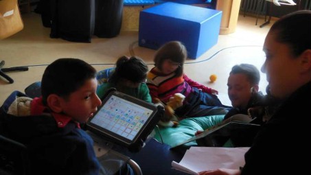 Das Foto zeigt eine Schülergruppe und zwei Erwachsene beim Bilderbuchlesen mit Buch und Talker.