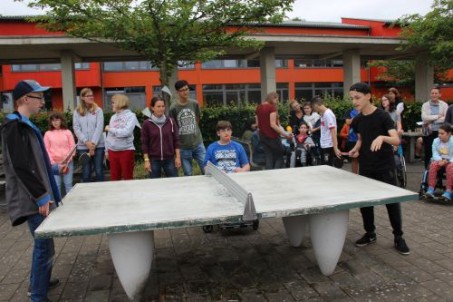 Schülerinnen und Schüler stehen um die Tischtennisplatte herum. Zwei Schüler spielen Tischtennis und ien Schüler im Rollstuhl ist Schiedsrichter.