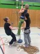 Eine Kollegin klettert an einem in den Ringen eingehängten Seil hoch gesichert durch zwei andere Kolleginnen, die alle Klettergurte an haben.
