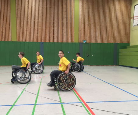 Vier Schüler der Schulmannschaft stehen mit gelben Trikots in ihren Rollstühlen auf dem Basketballfeld in der Turnhalle in Startaufstellung bereit zum Spiel.