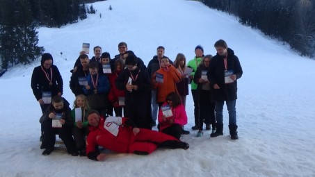 Gruppenfoto vor der beschneiten Piste mit Urkunden und Medaillen. Der Skilehrer liegt vor den Schülern im Schnee.